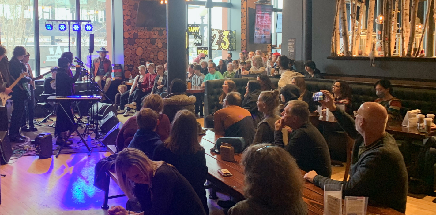 A packed audience watching an indie band perform on stage at MetroNOME Brewery, featuring guitarists, a keyboardist, and a vocalist.