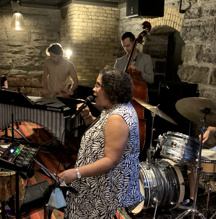 A jazz singer performing with a band at MetroNOME Brewery. The band features a vibraphonist, a double bassist, and a drummer.